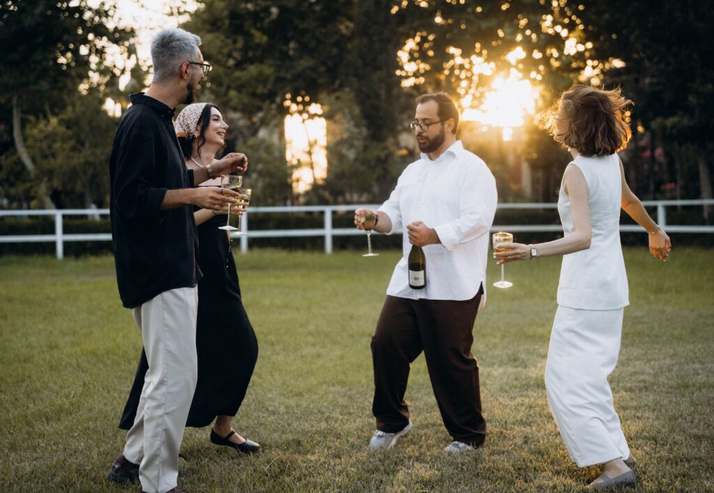 Four adults enjoying an outdoor celebration, dancing and holding drinks as the sun sets.